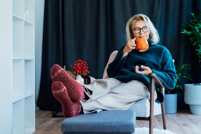 Middle aged woman relaxing with pumpkin shaped cup of hot drink in scandy style hygge interior home with fall mood decor. Lady dreaming, enjoy calm mood without stress, well being alone, self-care concept