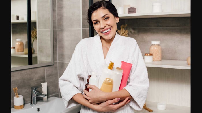 woman in robe in bathroom arms full of personal care bottles and products