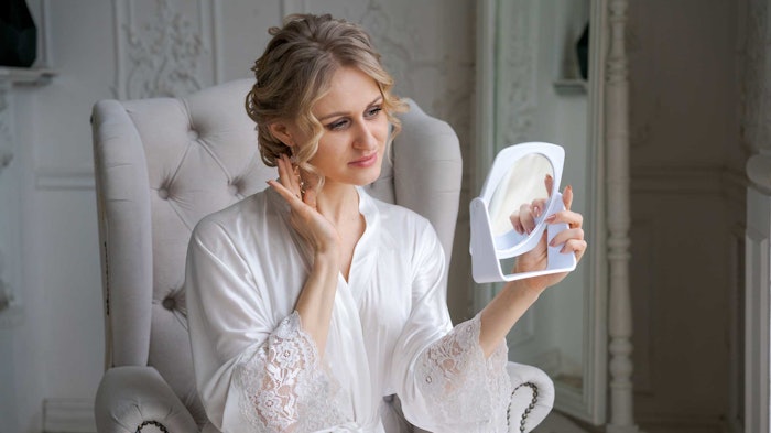caucasian woman in white silky dress in victorian chair admiring face in mirror