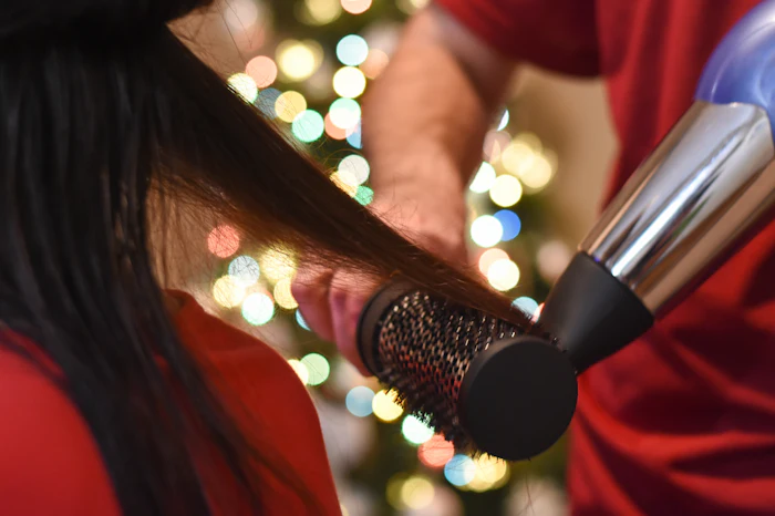 Hairdresser styling a client's hair with a blowdryer and round brush with a holiday tree in the background.