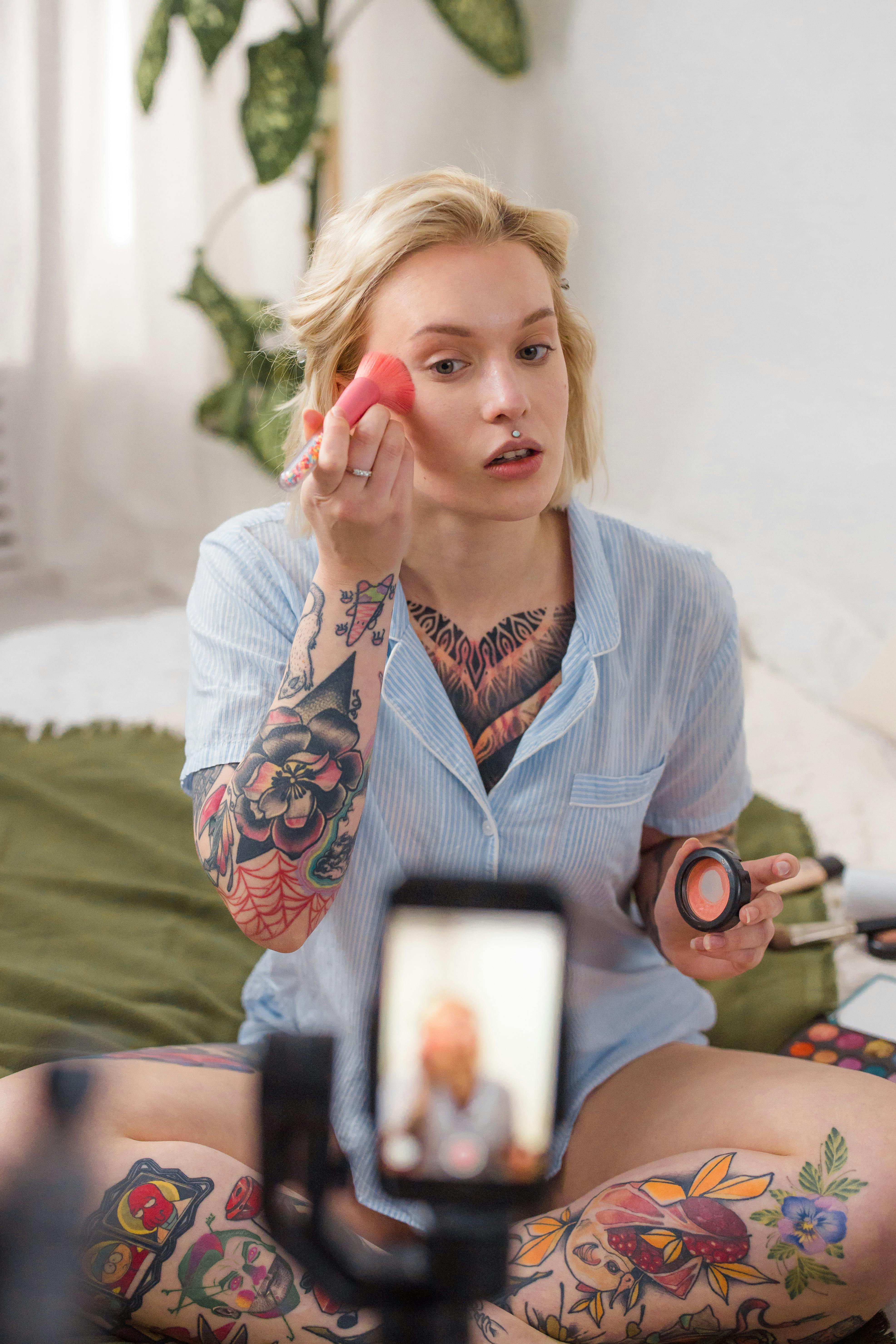 Tattooed woman applying makeup with beauty blender, filming tutorial on smartphone
