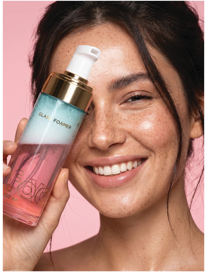 Woman holding GLAS FOAMER facial cleanser bottle, smiling against pink background with visible freckles