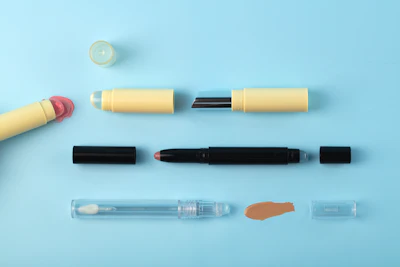 Assorted makeup products including lipsticks, eyeshadow applicators, and foundation swatches arranged on a light blue background