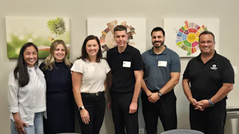 Six professionals smiling at camera in office setting with sustainability graphics on wall behind them