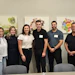 Six professionals smiling at camera in office setting with sustainability graphics on wall behind them