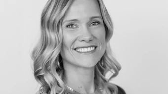 Professional headshot of smiling woman with wavy blonde hair wearing dark top and pearl necklace