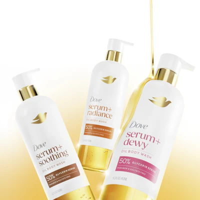 Three Dove serum+ oil body wash bottles with gold pumps displayed against a golden gradient background