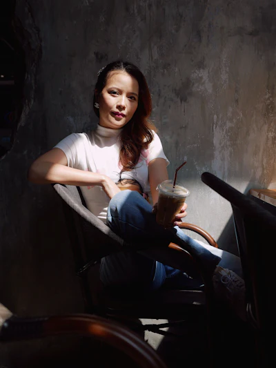 Woman in white t-shirt and jeans sitting against textured wall holding iced beverage
