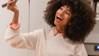 Woman with curly hair laughing while holding a whisk with chocolate batter dripping in a bright kitchen