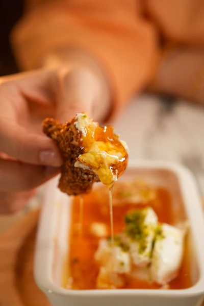 Hand holding crispy fried food with golden honey dripping over a white bowl of sauce with herbs