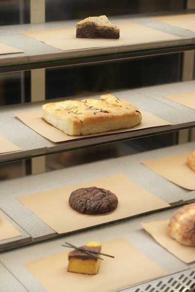 Tiered bakery display case showcasing assorted baked goods including focaccia, cookies, brownies, and pastries on parchment paper