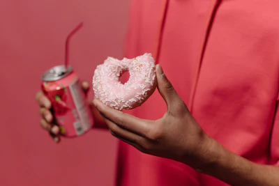 Person in pink shirt holding heart-shaped frosted donut and beverage can against pink background