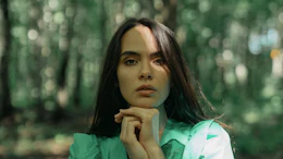 Woman in green jacket standing thoughtfully in a sunlit forest with trees in soft focus behind her