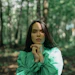 Woman in green jacket standing thoughtfully in a sunlit forest with trees in soft focus behind her
