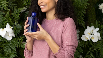 Woman in mauve sweater holding blue cosmetic bottle against green foliage and white flowers