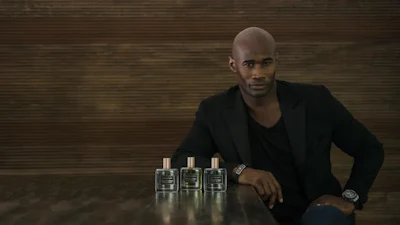 Professional man in black blazer posing with three fragrance bottles on a wooden surface