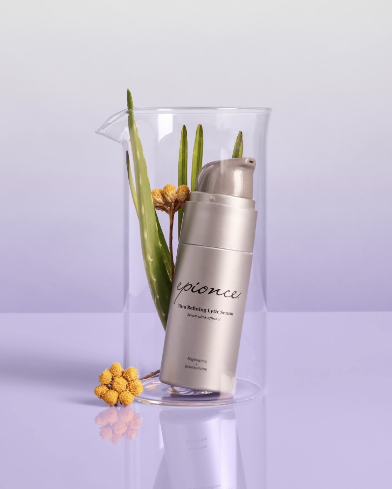 Epionce Ultra Refining Lytic Serum bottle displayed in clear cylinder with green plant stems and yellow flowers on lavender background