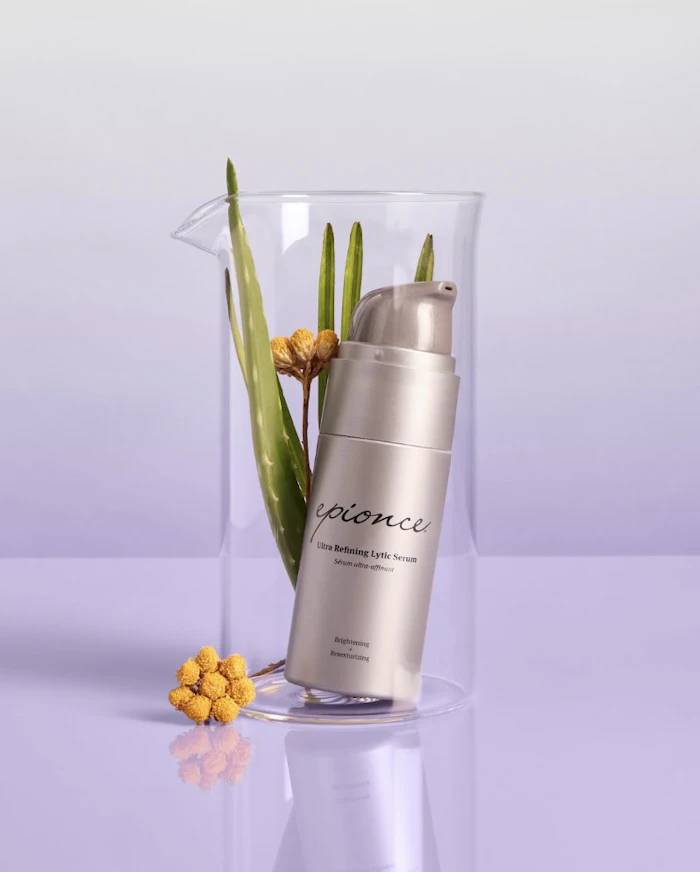 Epionce Ultra Refining Lytic Serum bottle displayed in clear cylinder with green plant stems and yellow flowers on lavender background