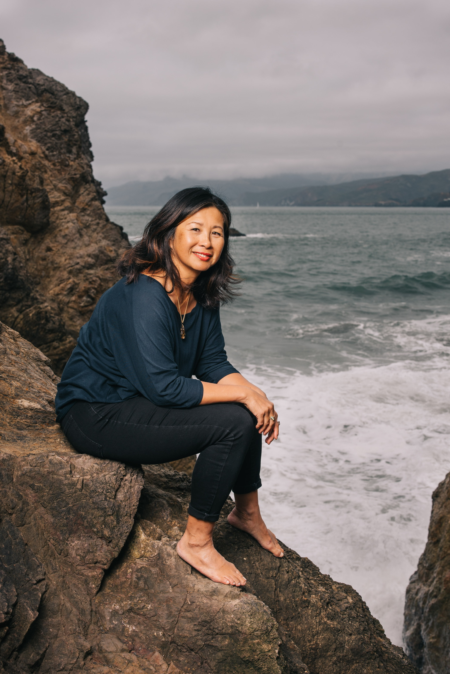 Woman smiling while sitting on rocky outcrop overlooking ocean waves and distant coastline