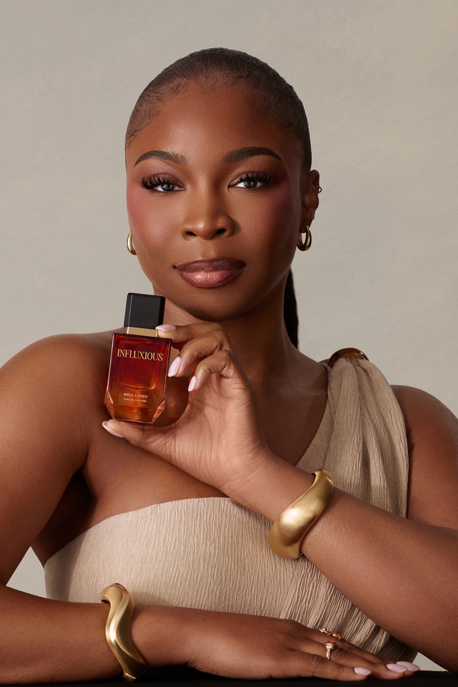 Woman holding Influxious fragrance bottle wearing gold bracelets and neutral draped clothing