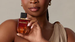Woman holding Influxious fragrance bottle wearing gold bracelets and neutral draped clothing