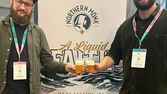 Two men with event badges stand beside a Northern Monk brewery collaboration poster for A Liquid Fate Hazy Session IPA beer