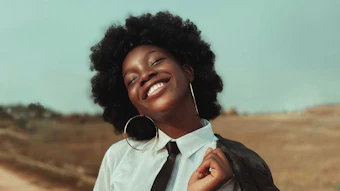 Smiling woman in white shirt and black tie with hoop earrings standing in desert landscape under blue sky