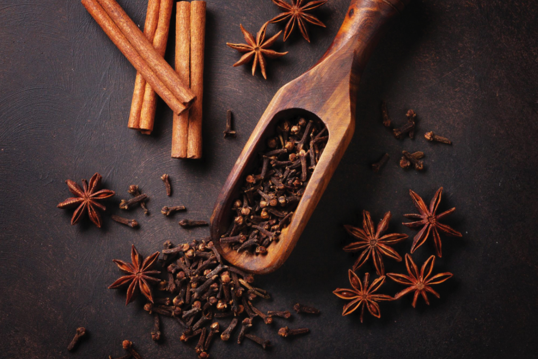 Cinnamon sticks, star anise, and cloves arranged with wooden scoop on dark surface