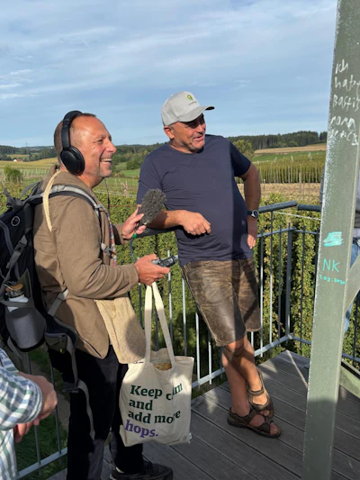 Two men conversing at a vineyard overlook, one wearing headphones holding a tote bag with hop-related text