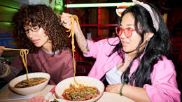 Two people eating noodles at a vibrant neon-lit restaurant, enjoying Asian cuisine together