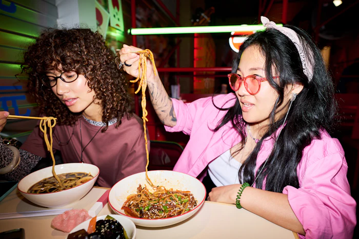 Two people eating noodles at a vibrant neon-lit restaurant, enjoying Asian cuisine together
