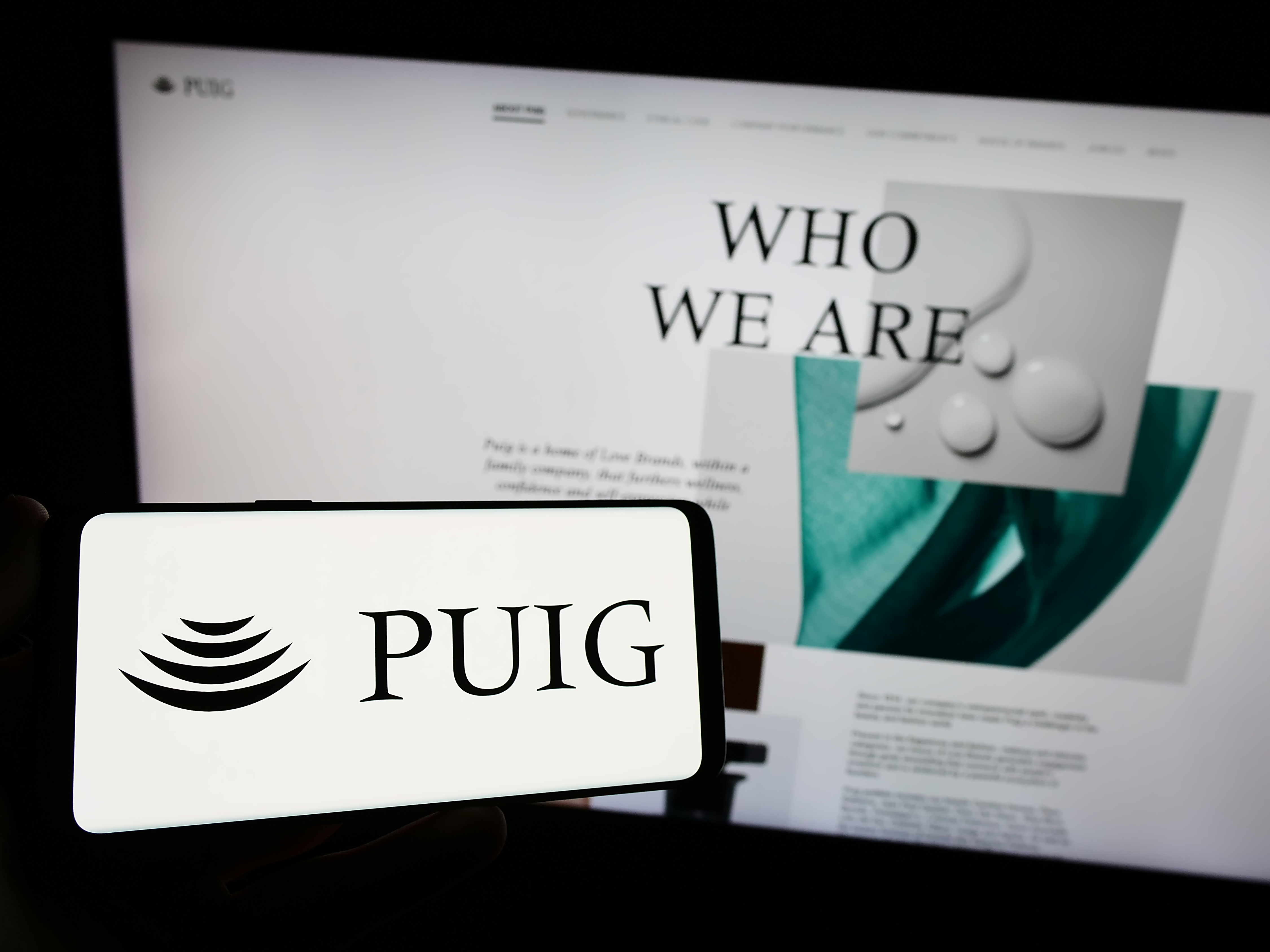 PUIG logo displayed on smartphone screen with 'Who We Are' website content visible in background