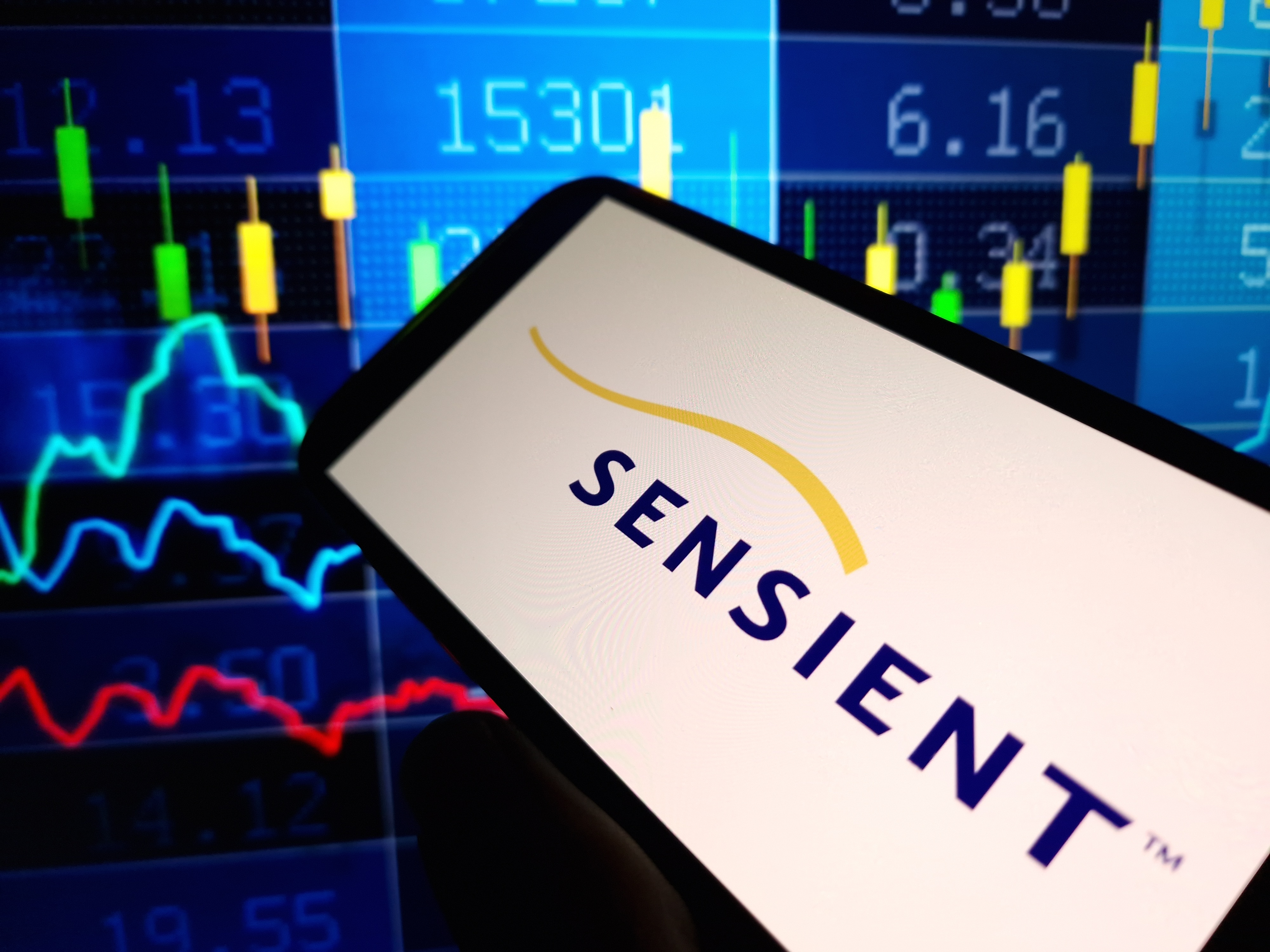 Sensient logo displayed on smartphone screen with financial trading charts and market data in background