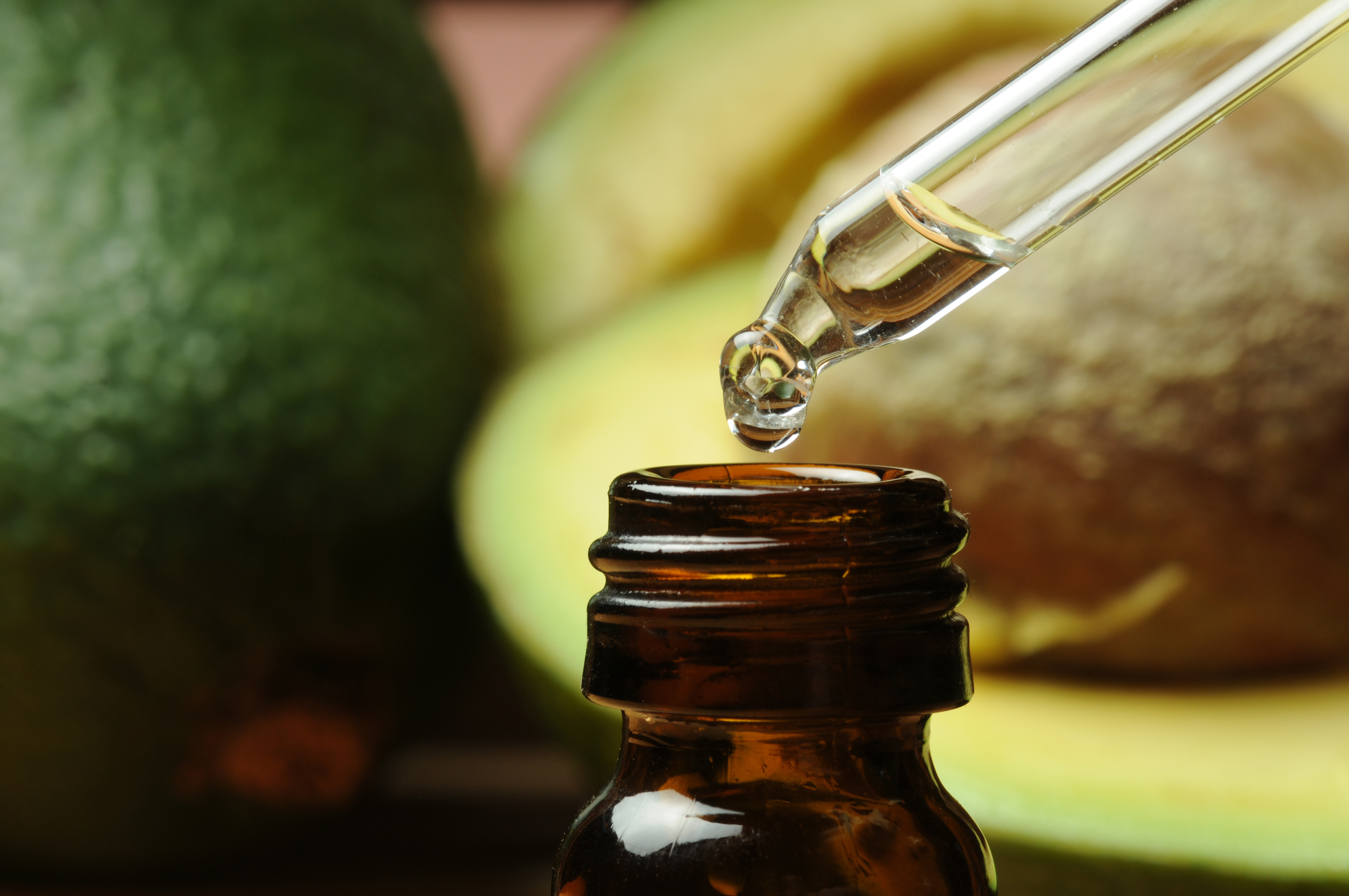 Golden essential oil or serum being dispensed from a dropper into a dark amber glass bottle with fresh avocados in the background