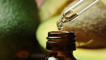 Golden essential oil or serum being dispensed from a dropper into a dark amber glass bottle with fresh avocados in the background