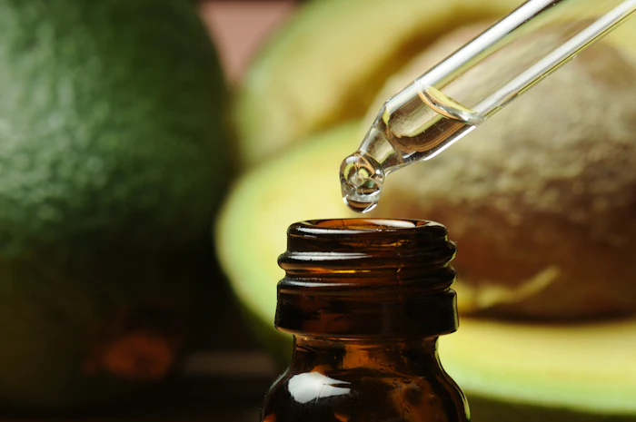 Golden essential oil or serum being dispensed from a dropper into a dark amber glass bottle with fresh avocados in the background