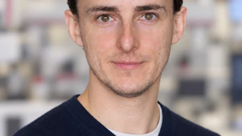 Professional headshot of a man with dark hair wearing a navy sweater in an office setting