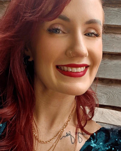 Smiling woman with burgundy wavy hair, red lipstick, nose ring, and gold chain necklace against wooden background
