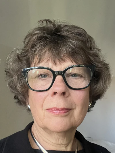 Professional headshot of a woman wearing black-framed glasses and black top against neutral background