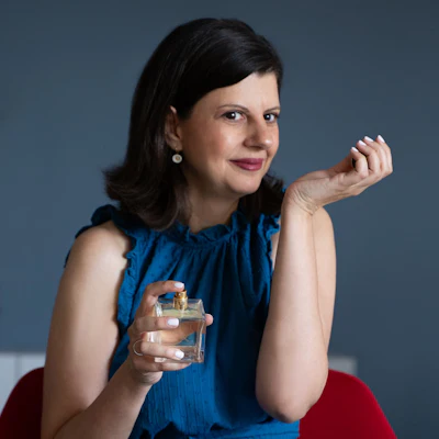 Professional woman in blue sleeveless top holding perfume bottle against gray background