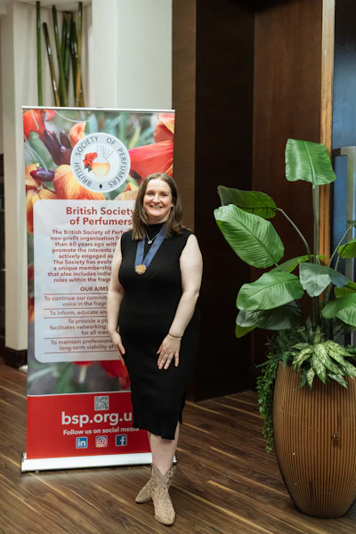 Woman in black dress standing beside British Society of Perfumers promotional banner with floral design and organizational information
