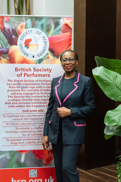 Professional woman in navy pinstripe blazer with pink trim standing before British Society of Perfumers banner with floral background