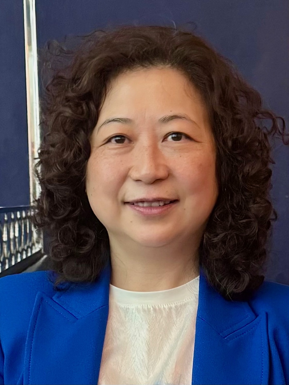 Professional headshot of a woman with curly dark hair wearing a bright blue blazer and white top against a dark blue background