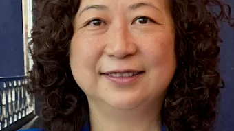 Professional headshot of a woman with curly dark hair wearing a bright blue blazer and white top against a dark blue background