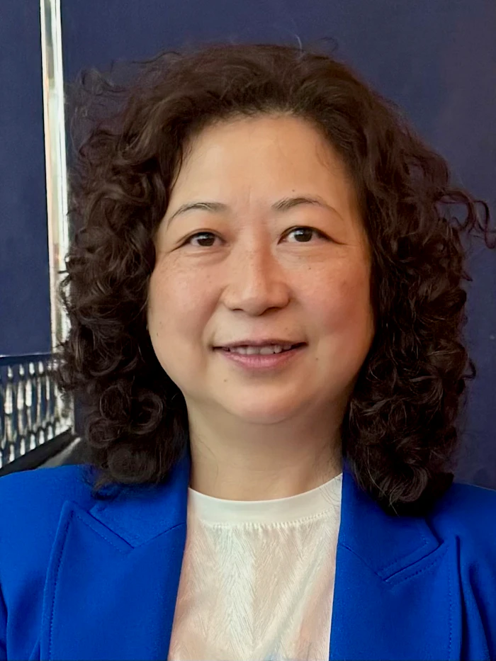 Professional headshot of a woman with curly dark hair wearing a bright blue blazer and white top against a dark blue background