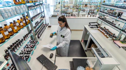 Scientist in lab coat working at bench with shelves of bottles and products in modern laboratory