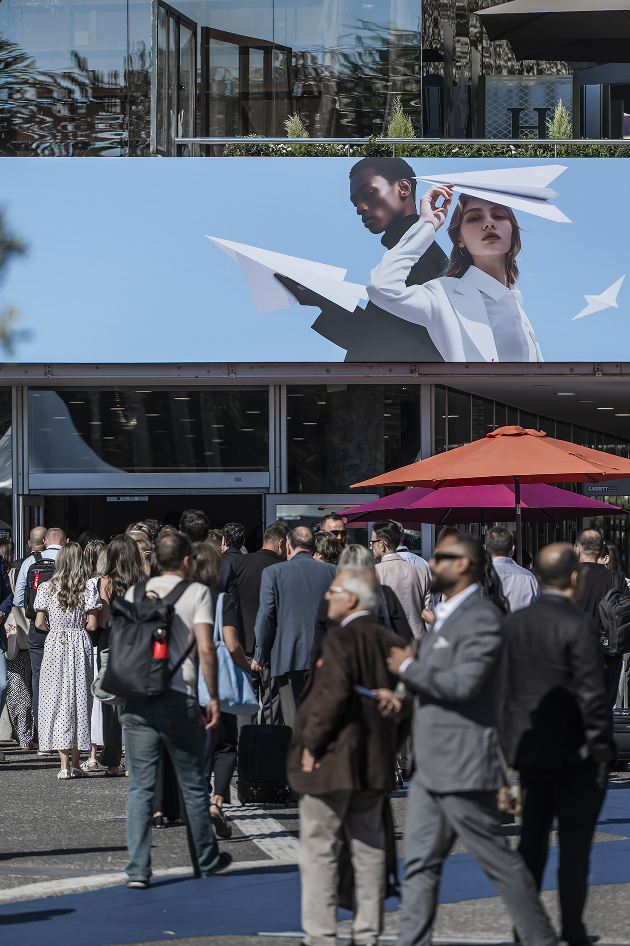 Crowd gathered at outdoor event beneath large billboard featuring two people with paper airplanes against blue sky