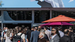 Crowd gathered at outdoor event beneath large billboard featuring two people with paper airplanes against blue sky