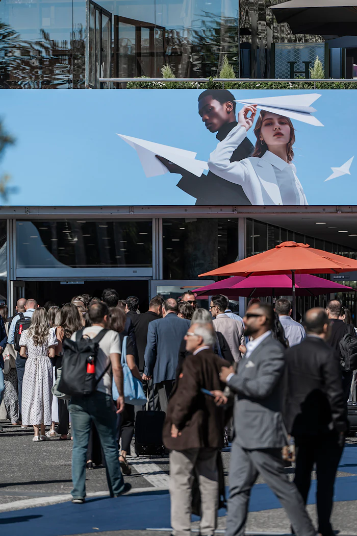 Crowd gathered at outdoor event beneath large billboard featuring two people with paper airplanes against blue sky