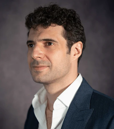 Professional headshot of a man in dark blazer and white shirt against neutral gray background